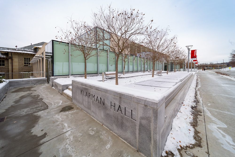 The glass facade of a long, low contemporary building shown in winter with a planter in the foreground containing trees and the words "Klarman Hall"
