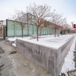 The glass facade of a long, low contemporary building shown in winter with a planter in the foreground containing trees and the words "Klarman Hall"