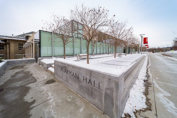 The glass facade of a long, low contemporary building shown in winter with a planter in the foreground containing trees and the words "Klarman Hall"