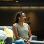 A Black woman in a light green top and leans sits on a couch, talking with several other people, who are mostly out of frame.