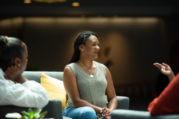 A Black woman in a light green top and leans sits on a couch, talking with several other people, who are mostly out of frame.
