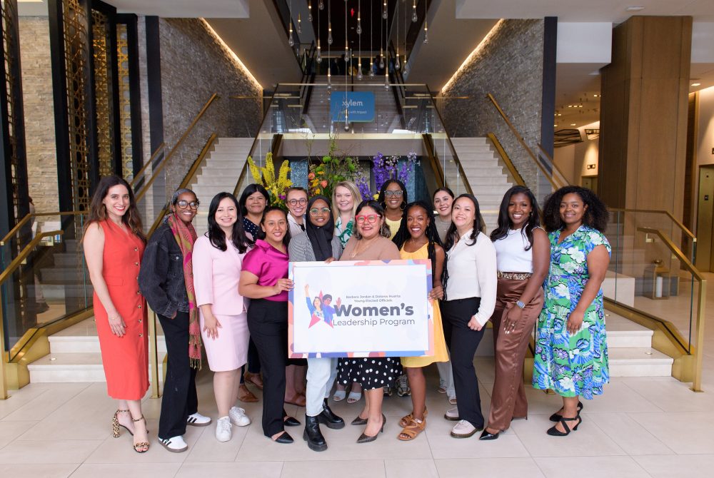 A group of women standing in a conference center or hotel lobby holding a sign.