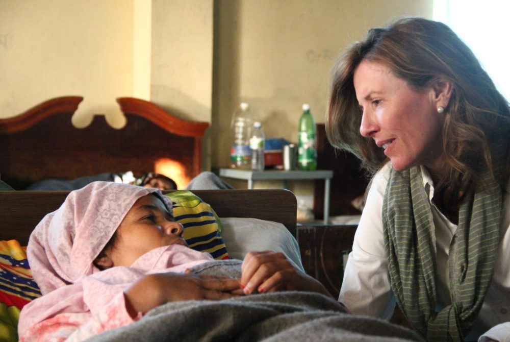 Article Banner - A woman wearing a scarf (Kate Grant) looks at another woman lying in bed.