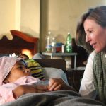 Article Banner - A woman wearing a scarf (Kate Grant) looks at another woman lying in bed.