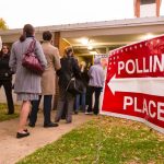A red sign in the foreground points a line of voters toward a polling place.