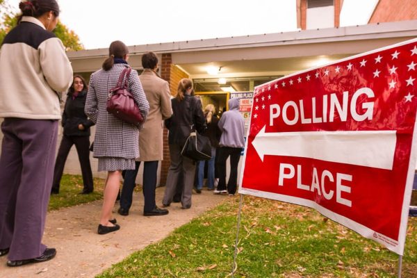 A red sign in the foreground points a line of voters toward a polling place.