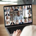 A man looks at a laptop featuring a gallerry-style Zoom screen for a digital meeting.