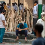 Article Banner - Men prepare funeral pyres for COVID patients in New Delhi, India, while one man sits on the curb with his head down.