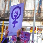 A woman holds up a sign depicting a raised fist inside the "woman" symbol, at a protest.