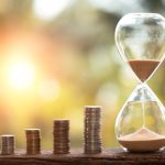 Piles of stacked coins next to an hourglass against a blurred sunset background.