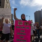 A young woman wearing a hijab holds a sign opposing guns and an abortion ban, flanked by other protesters.