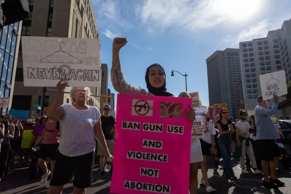 A young woman wearing a hijab holds a sign opposing guns and an abortion ban, flanked by other protesters.