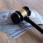 Article Banner - Gavel lying atop a stack of hundred-dollar bills, on a wooden tabletop.
