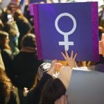 Article Banner - Women protest holding sign with female symbol in white against a purple background