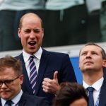 A man in a suit (Prince William) gives a thumbs-up sign while smiling, standing amid several other men in suits.