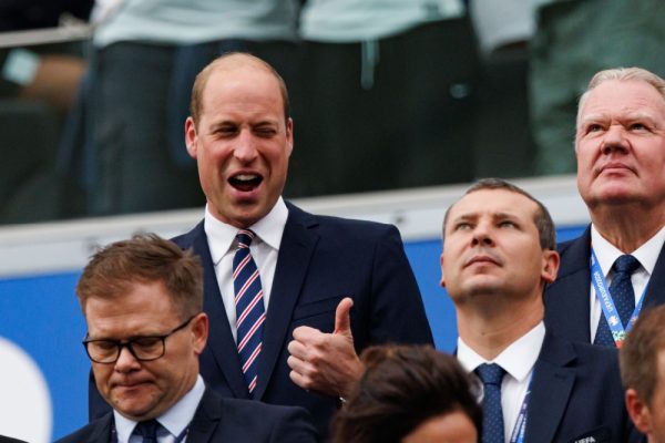 A man in a suit (Prince William) gives a thumbs-up sign while smiling, standing amid several other men in suits.