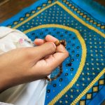 Close-up of woman's hand holding Muslim prayer beads, against a blue and yellow prayer mat or carpet.