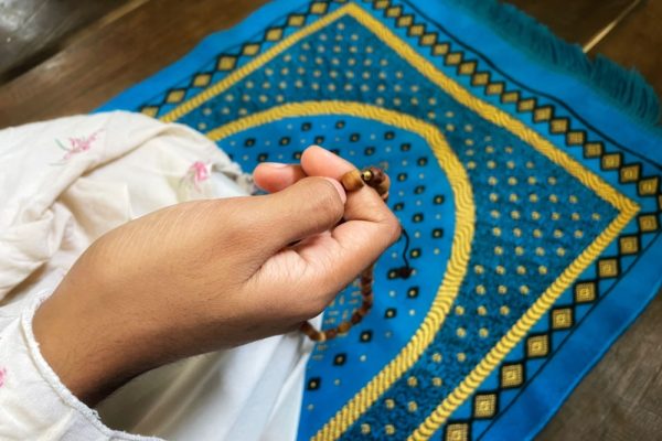 Close-up of woman's hand holding Muslim prayer beads, against a blue and yellow prayer mat or carpet.