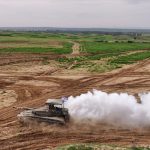 An Israeli tank moves over dusty ground with green fields in the background.
