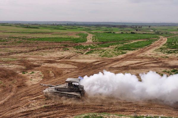 An Israeli tank moves over dusty ground with green fields in the background.
