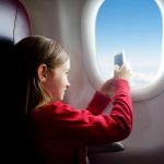 Girl in red shirt taking a phone picture while looking at the sky through an airplane window.