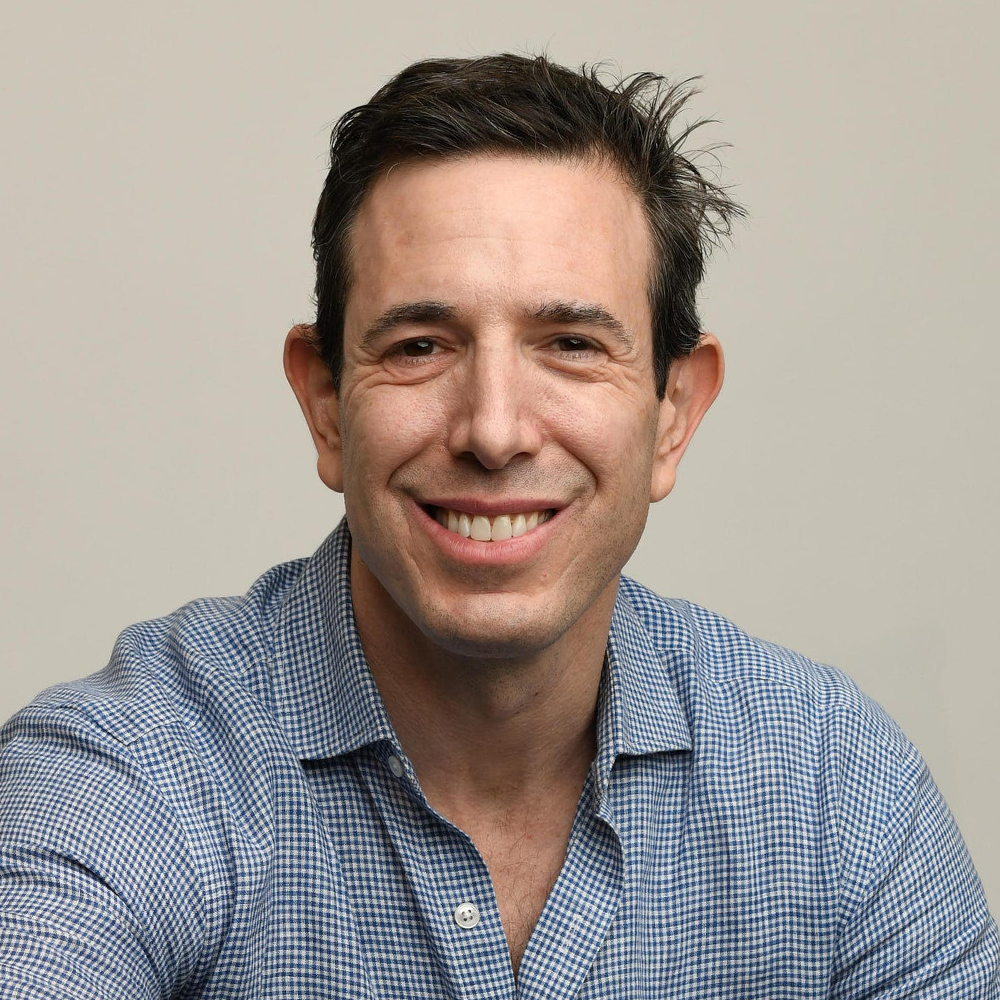 Headshot of a smiling middle-aged white man with dark brown hair wearing a light blue checked button-down shirt.